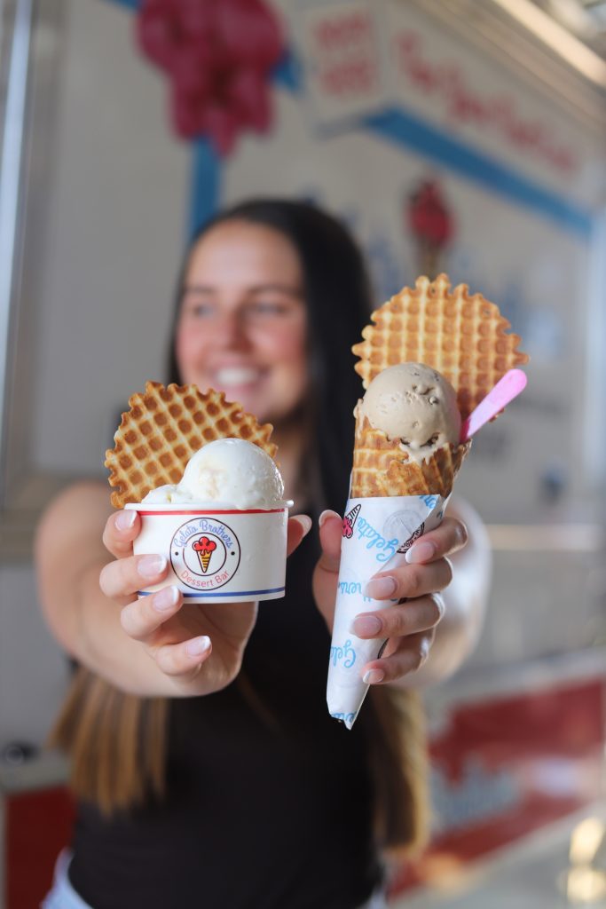 A smiling woman holds up two waffle cones topped with scoops of gelato in paper cups.