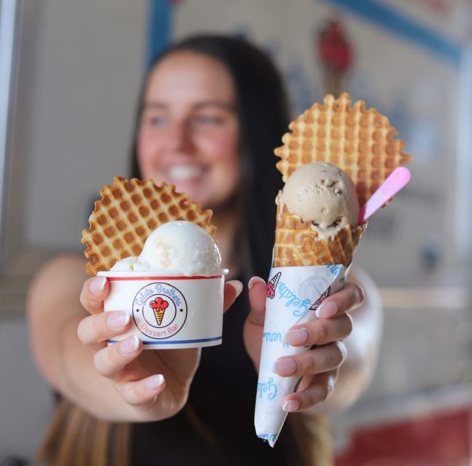 A smiling woman holding two waffle cones topped with scoops of gelato in branded cups.
