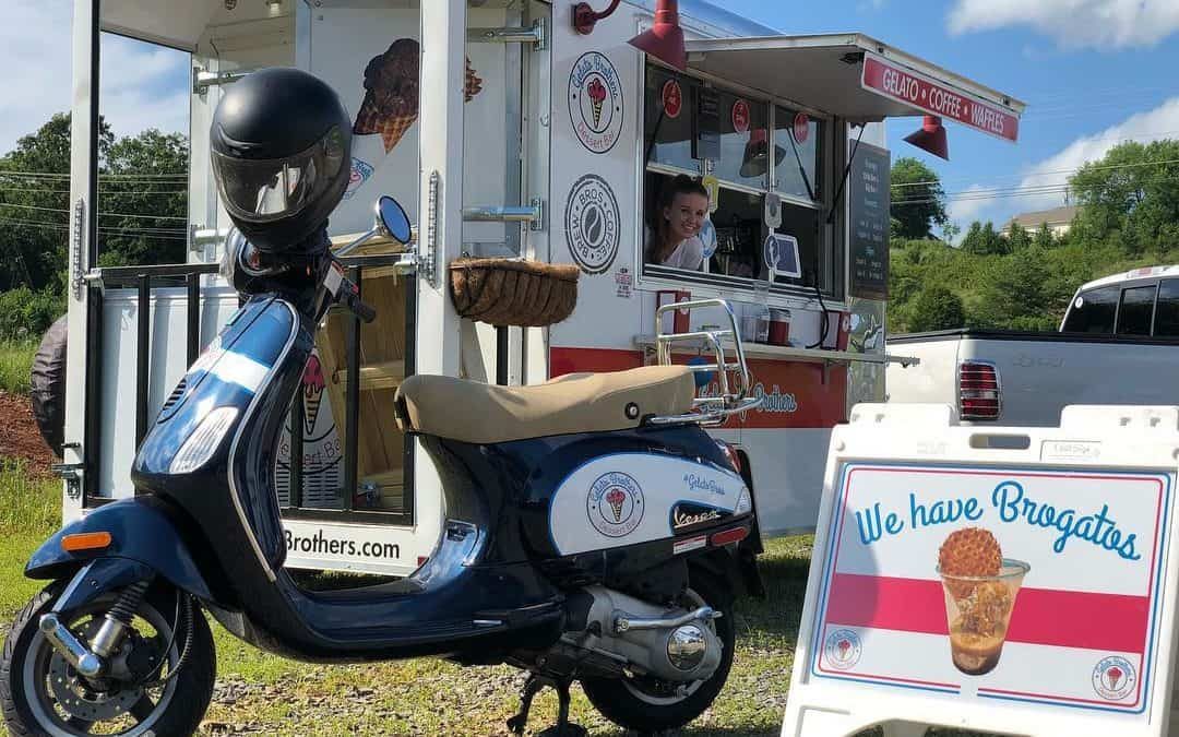 Blue and white Vespa scooter parked beside a vintage gelato cart trailer on a sunny day.
