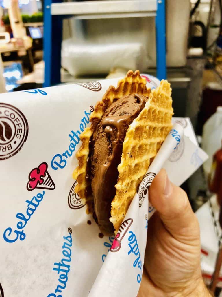 A hand holds a golden waffle cone sandwich filled with rich chocolate gelato wrapped in Gelato Brothers branding paper.