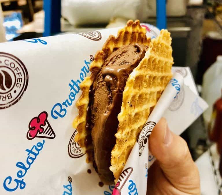 A hand holds a waffle cone sandwich filled with chocolate gelato and waffle cookie pieces wrapped in branded paper.