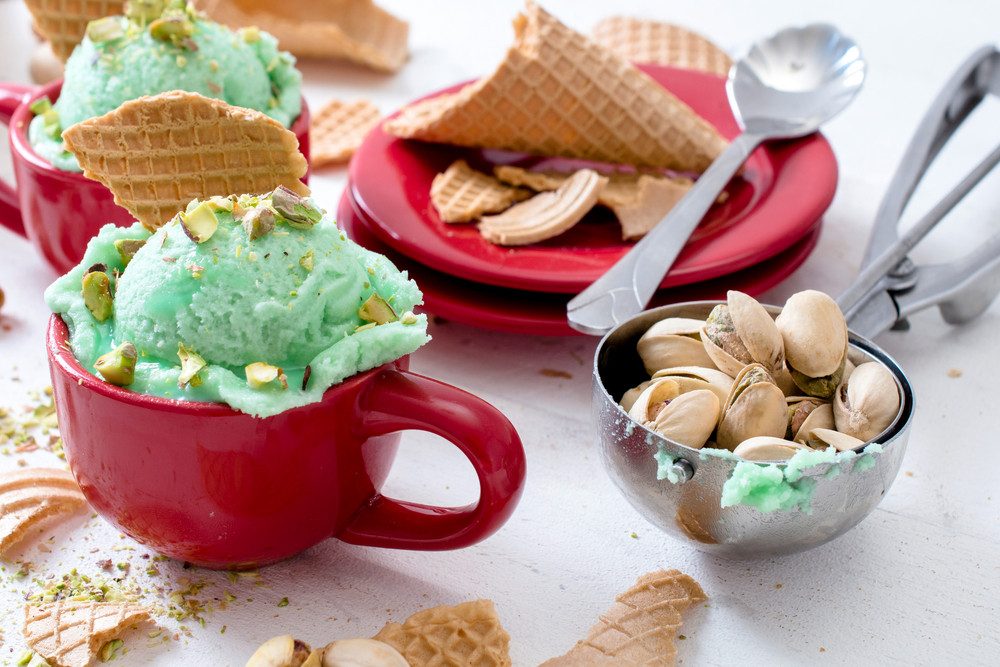 Pistachio gelato served in red mugs with waffle cones, pistachios, and cookies on a white surface.