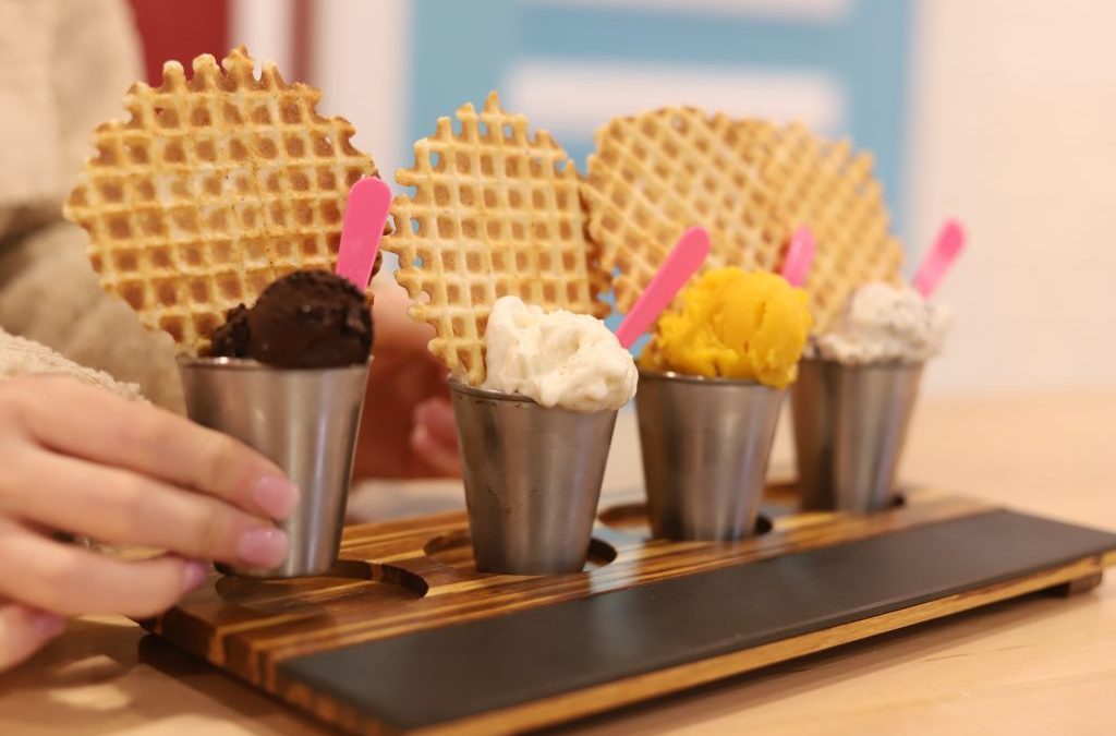 Four waffle cone gelato cups with colorful toppings arranged on a wooden paddle at a tasting event.