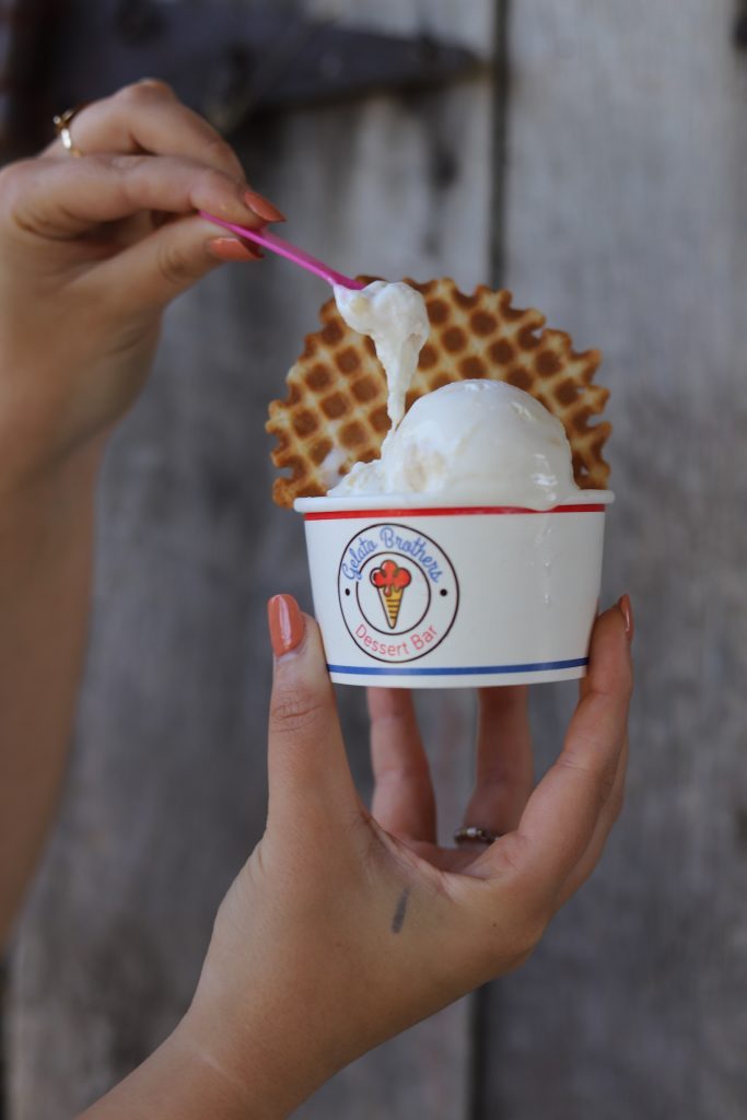 A hand holds a cup of vanilla gelato topped with a waffle cone against an urban background.