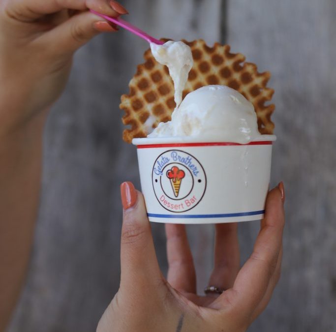 Hand holding a cup of vanilla gelato topped with a waffle cone and pink spoon.