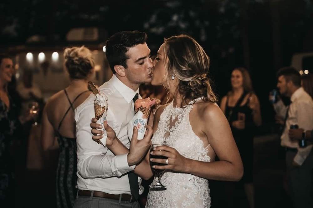 A couple in formal attire shares a kiss while holding champagne glasses at an evening celebration surrounded by guests.