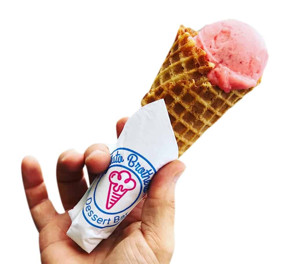 A hand holding a waffle cone wrapped in white paper with pink gelato scoops against a white background.