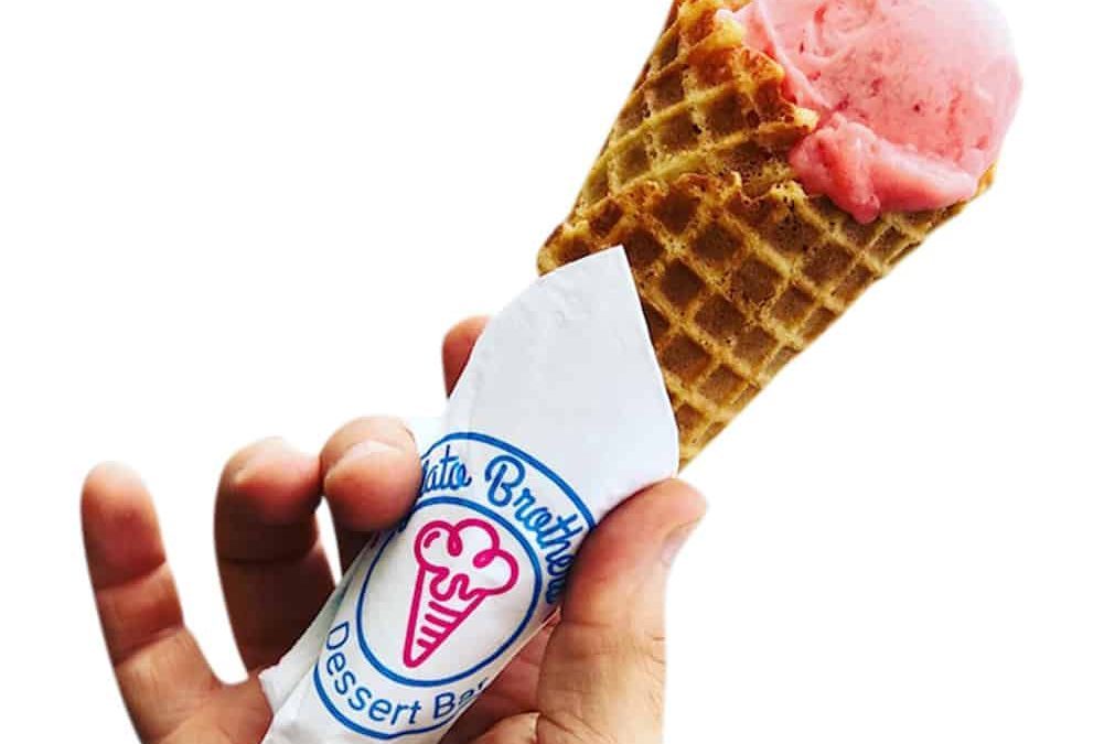 A hand holds a waffle cone with pink gelato wrapped in a branded napkin against a white background.