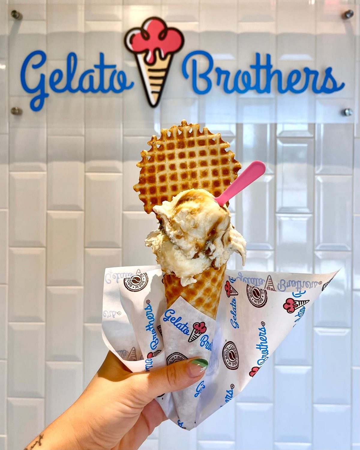 A hand holds a waffle cone topped with gelato and a pink spoon in front of the Gelato Brothers neon sign.