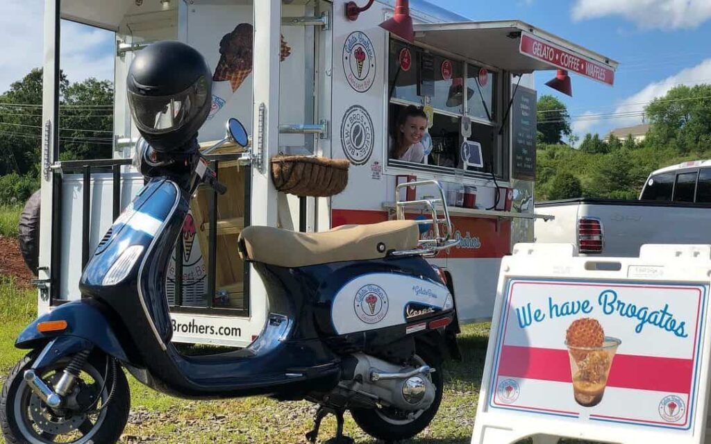 Blue and white Vespa scooter parked beside a red and white food trailer with serving window.