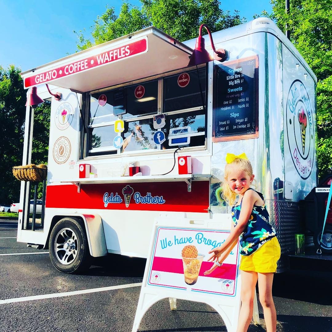 A young child stands beside a Gelato Brothers food trailer offering candy, coffee and waffles at an outdoor event.