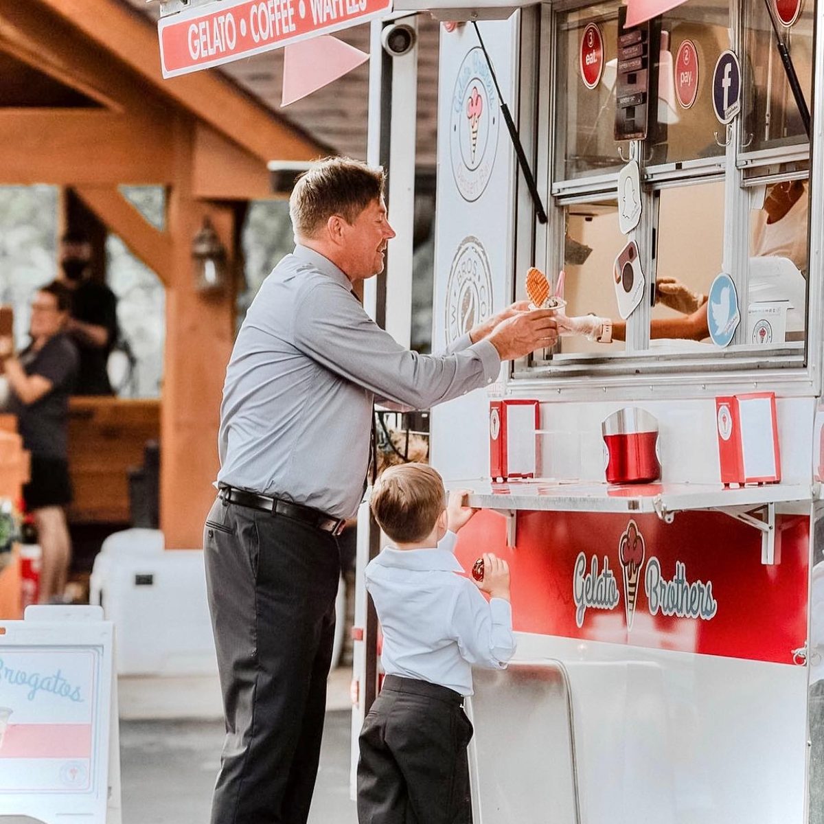 A man and young child ordering from a red and white gelato cart at an outdoor event.