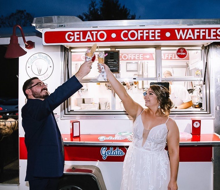 A couple in wedding attire toast with gelato cups in front of a red and white dessert cart at dusk.