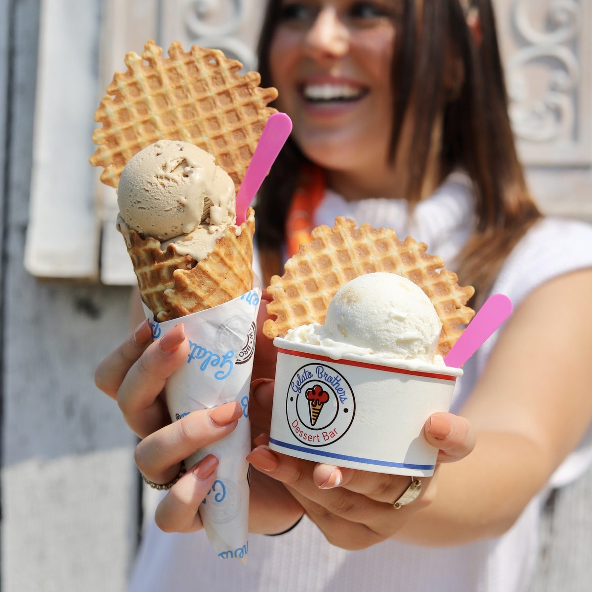 A smiling woman holds two waffle cones topped with vanilla gelato in branded cups outdoors.
