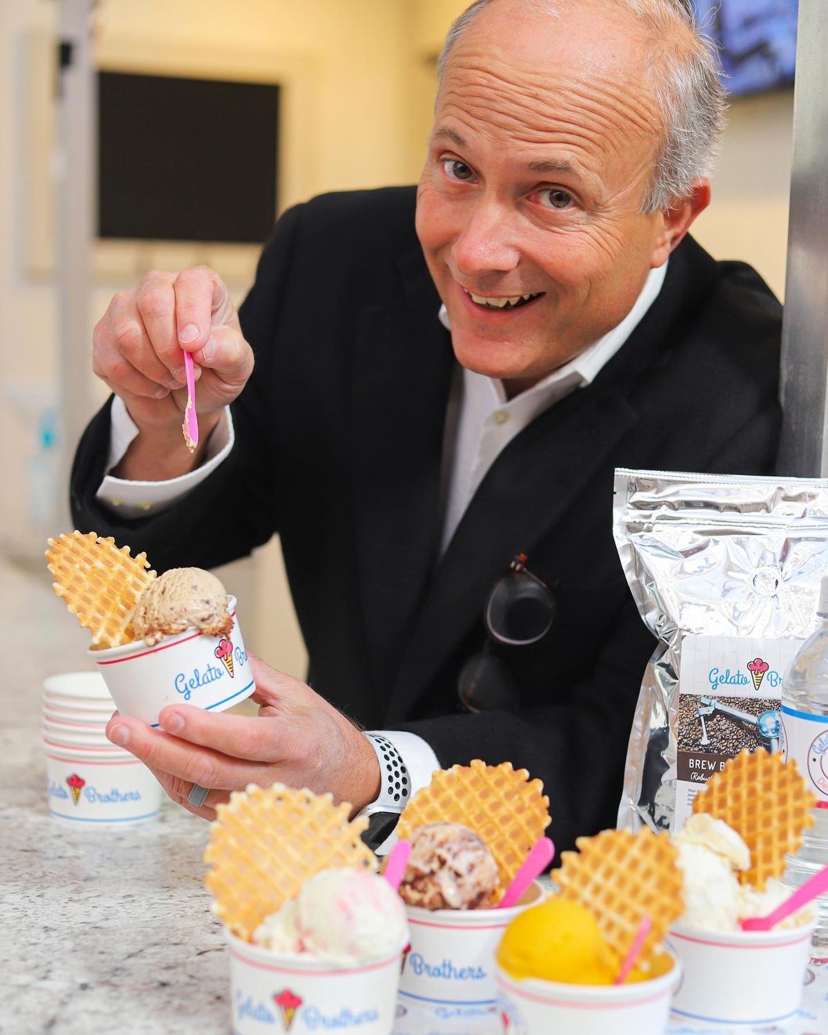 A man in a black blazer smiles while holding a spoon over a cup of gelato surrounded by other gelato cups.