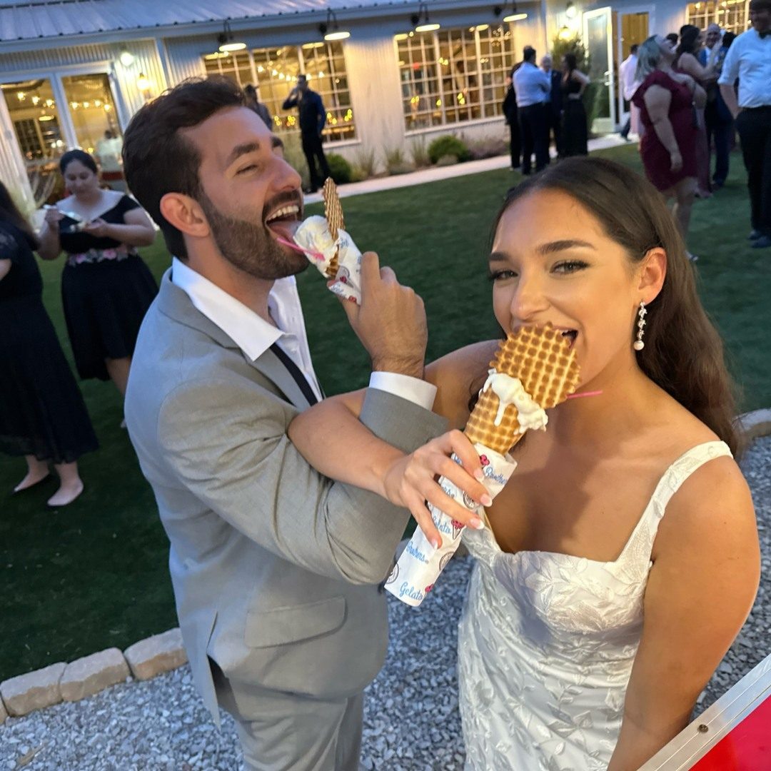 A couple in formal attire enjoys gelato together at an outdoor evening event on a lawn.
