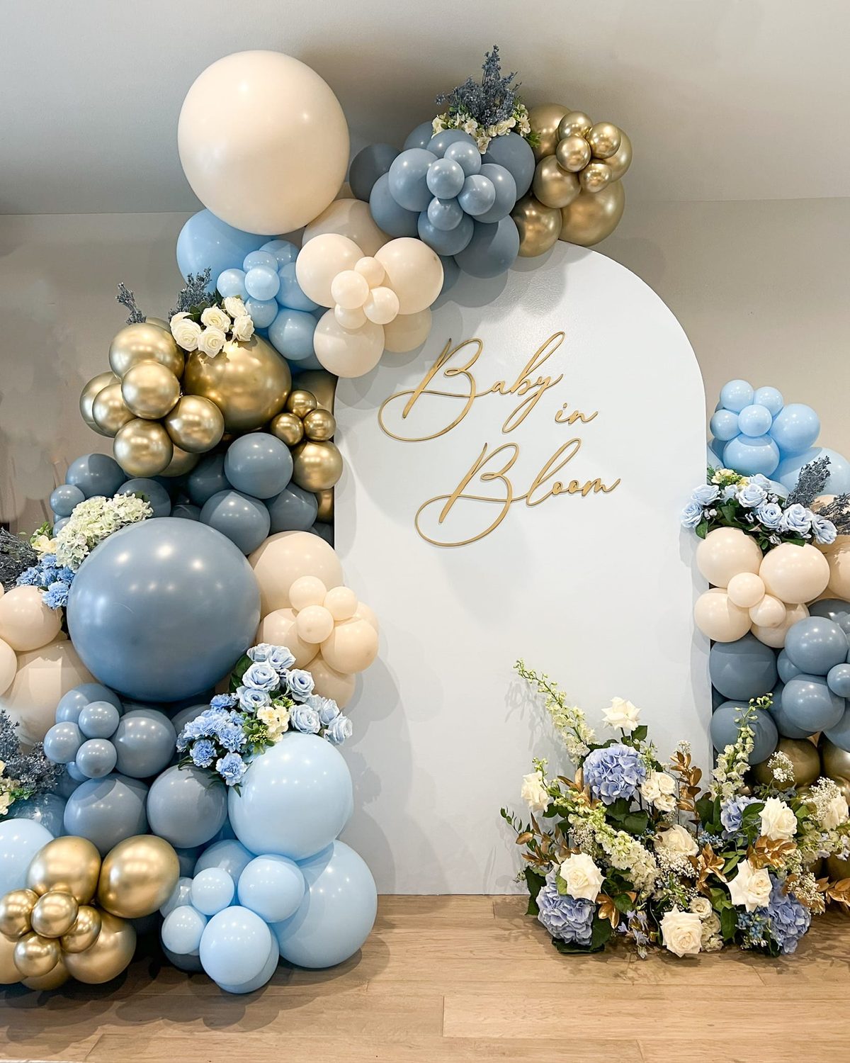 An elegant white arch decorated with blue, cream, and gold balloons and white flowers displays "Baby in Bloom" text.