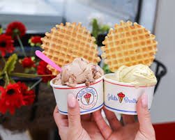Two cups of gelato topped with waffle cookies held in front of a pink gelato cart.
