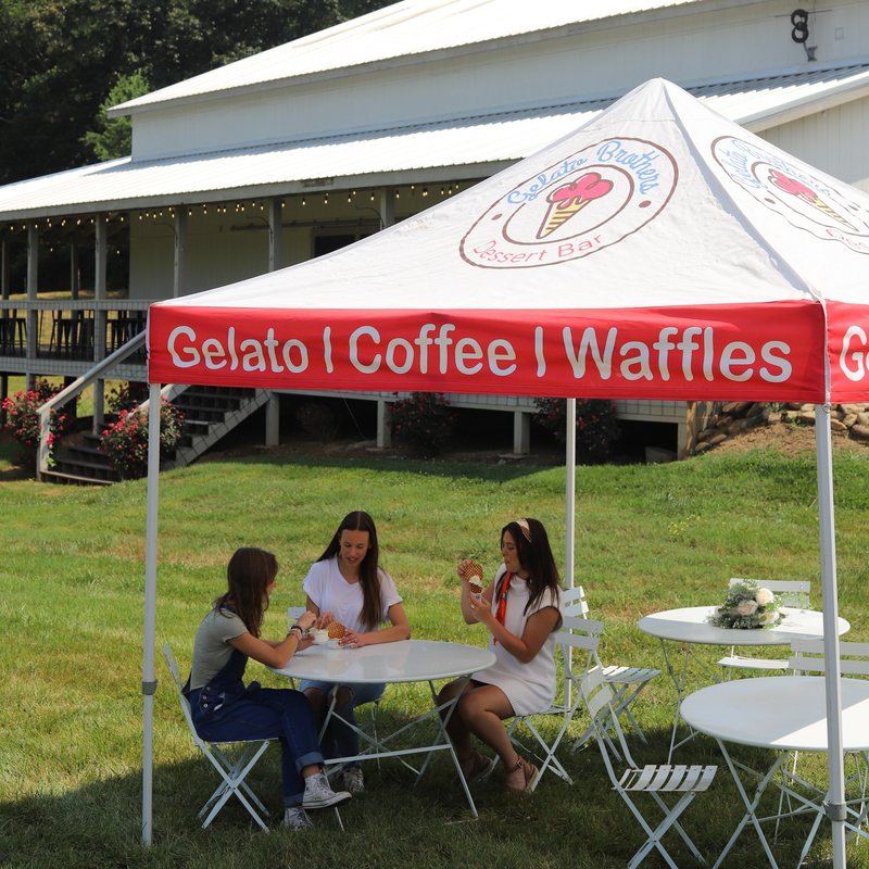 Guests with gelato at white bistro tables and chairs under a Gelato Brothers tent, barn with string lights behind